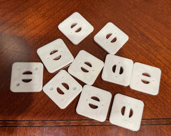 May include: A collection of ten white square plastic buttons, each featuring rounded corners and a central horizontal slot flanked by two small circular holes. The buttons are arranged in a scattered pattern, displaying a smooth, flat surface.