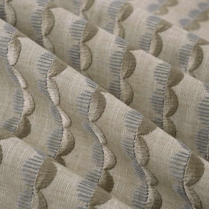 May include: Close-up of a beige fabric with a repeating pattern of embroidered, scalloped shapes. The pattern alternates between light blue and taupe, creating a textured, layered effect. The fabric is folded, showing the depth and drape of the material.