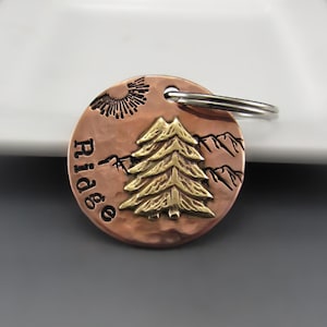 May include: Copper dog tag with a gold-colored pine tree design and the word "Ridge" engraved on it. The tag has a silver key ring attached.