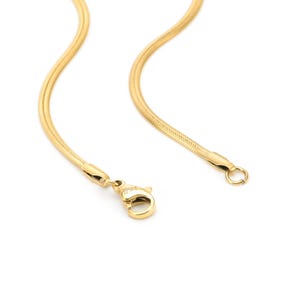 May include: A gold-coloured snake chain necklace with a lobster clasp and a small ring at the end. The necklace is made of small, interlocking links that create a smooth, flexible chain. The background is white.