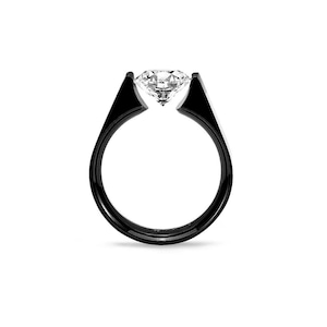 Pode incluir: Um anel com uma banda preta lisa e polida com um engaste de tensão que segura uma única pedra preciosa transparente de corte redondo no topo. O design cria um espaço entre as duas extremidades da banda onde a pedra está suspensa.