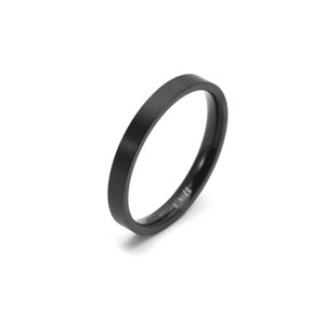 Könnte beinhalten: Ein schwarzer Ring mit mattem Finish. Der Ring hat ein schlichtes Banddesign mit flacher Oberseite und abgerundeten Kanten. Der Ring ist vor einem weißen Hintergrund platziert.