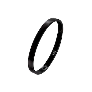 Puede incluir: Una pulsera rígida de color negro sólido con un acabado liso y pulido. La pulsera cuenta con un mecanismo de cierre de bisagra e incluye el texto "b.tiff" impreso en una tipografía cursiva blanca en la superficie interior de la banda.