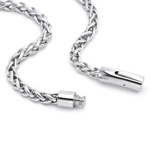 Puede incluir: Primer plano de un collar de cadena plateada con un patrón de trigo trenzado. El collar tiene un cierre seguro. La joya está hecha de un material metálico brillante.