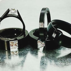 Pode incluir: Uma cole&ccedil;&atilde;o de an&eacute;is pretos com v&aacute;rios designs, incluindo bandas com pedras claras quadradas incrustadas, um anel solit&aacute;rio com uma pedra redonda cravada e uma banda fina acentuada com uma s&eacute;rie de pequenas pedras claras redondas.