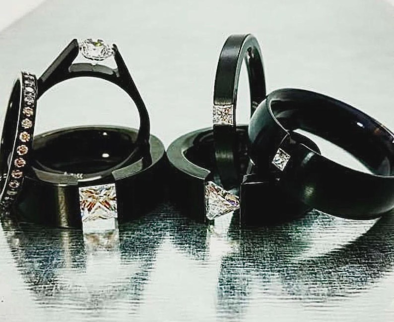 Pode incluir: Uma cole&ccedil;&atilde;o de an&eacute;is pretos com v&aacute;rios designs, incluindo bandas com pedras claras quadradas incrustadas, um anel solit&aacute;rio com uma pedra redonda cravada e uma banda fina acentuada com uma s&eacute;rie de pequenas pedras claras redondas.