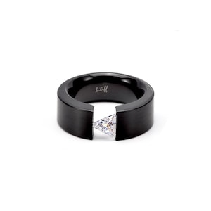 Pode incluir: Um anel preto com acabamento fosco, apresentando uma pedra preciosa triangular transparente fixada por tens&atilde;o na abertura central. O interior da banda est&aacute; gravado com o texto b.tiff. O anel tem um perfil largo e plano.