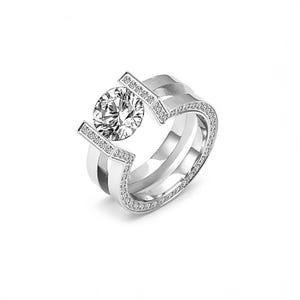 May include: A silver ring with a large, round, clear gemstone as the focal point. The ring has a unique design with two parallel bands and a decorative top section with small, embedded stones. The ring is set against a white background.