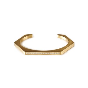 May include: A gold-toned, open cuff bracelet with a geometric design. The bracelet features a row of small, clear gemstones set along the flat front surface. The bracelet has a modern, minimalist aesthetic.