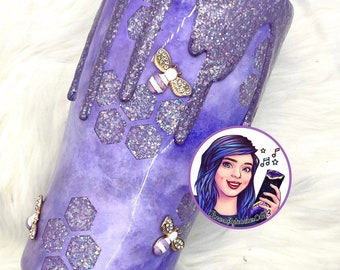 May include: A purple tumbler with a glittery top and dripping design. The tumbler features a honeycomb pattern with glitter accents and bee embellishments. A cartoon image of a woman with blue hair is on the side.