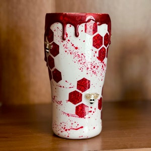 May include: White tumbler with a red honeycomb pattern and red dripping paint. The tumbler has small gold bee embellishments.