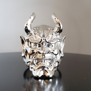 May include: A silver ring with a detailed design of a demon's face. The ring has two horns and a menacing expression.