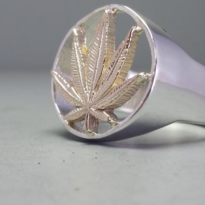 May include: A silver ring with a gold marijuana leaf design. The leaf is cut out of the ring, creating a negative space effect.
