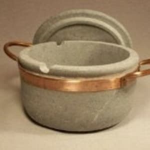 May include: A gray stone ashtray with a copper band and handles. The ashtray has a lid and a groove for resting cigarettes. The design is simple and functional, suitable for indoor or outdoor use.