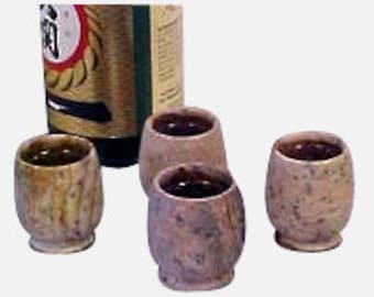 Puede incluir: Conjunto de cuatro pequeñas tazas de sake de cerámica moteada en marrón y blanco con una botella de sake en el fondo.