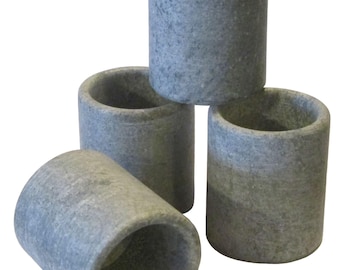 May include: Four gray, cylindrical stone candle holders. The holders are stacked in a pyramid shape, with one lying on its side. The stone has a speckled texture and a natural, earthy appearance.