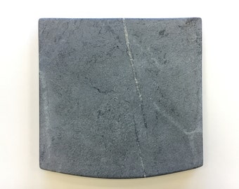 Puede incluir: Una tabla de servir de pizarra gris con un borde ligeramente curvado. La tabla tiene una textura natural y rugosa y una veta gris clara que la atraviesa.