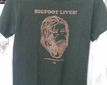 Puede incluir: Una camiseta verde oscuro con un gráfico marrón de Bigfoot y el texto "BIGFOOT LIVES!" impreso en la parte delantera.