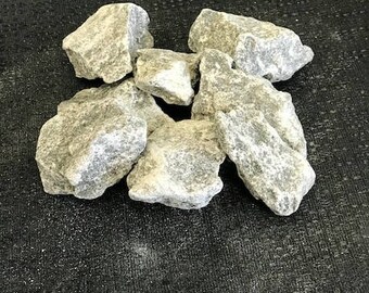 Puede incluir: Un conjunto de rocas de color gris claro, de forma irregular y con textura moteada. Las rocas están apiladas sobre una superficie oscura y texturizada, creando una estética natural y terrosa.