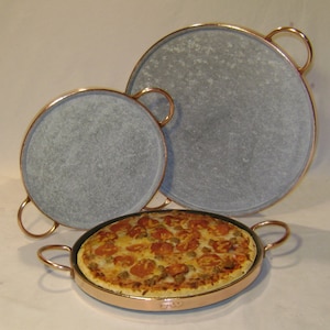 May include: Three round, gray stone pizza stones with copper rims and handles. The smallest stone has a pizza with red sauce, cheese, and toppings on it.