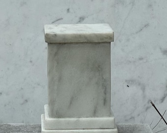 Puede incluir: Un pequeño pedestal cuadrado de mármol con una parte superior rectangular y una base escalonada. La piedra blanca tiene sutiles vetas grises. El pedestal está diseñado para exhibir objetos pequeños y añade un toque de elegancia.