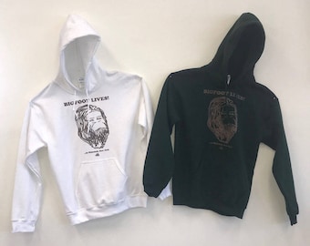 Puede incluir: Dos sudaderas con capucha con un gráfico de Bigfoot. La sudadera blanca tiene el texto "Bigfoot Lives!" y la sudadera verde tiene el texto "Bigfoot Lives!" Ambas sudaderas tienen un corte holgado.