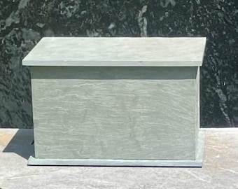 Puede incluir: Una caja rectangular de piedra gris claro con una parte superior inclinada. La caja tiene una superficie texturizada y una base. El fondo es una piedra verde oscuro y negra.