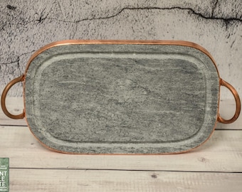 Puede incluir: Una bandeja rectangular de pizarra con asas de color cobre. La bandeja está hecha de piedra natural y tiene una superficie lisa y pulida.