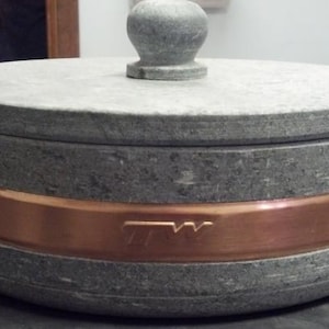 May include: A round, gray stone pot with a copper band around the rim and a knob on the lid. The pot has the letters "TW" embossed on the copper band.