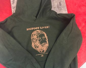 Puede incluir: Una sudadera con capucha verde oscuro con el texto "BIGFOOT LIVES!" en letras marrones. Debajo del texto hay un dibujo lineal marrón de un Bigfoot. El texto "In Whitehall, New York" está debajo del dibujo.