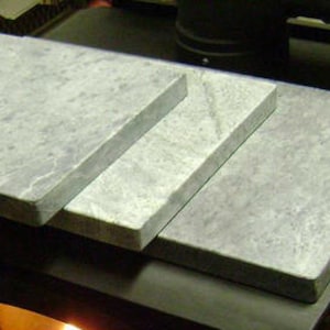 May include: Three rectangular gray soapstone tiles stacked on top of each other on a black surface. The tiles are smooth and have a light gray color.