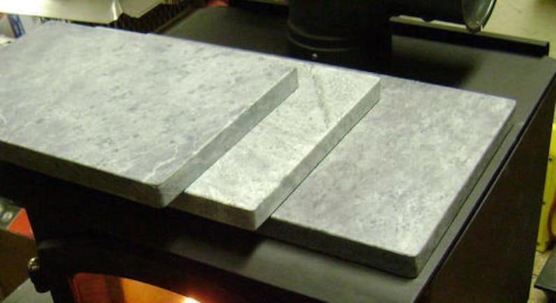May include: Three rectangular gray soapstone tiles stacked on top of each other on a black surface. The tiles are smooth and have a light gray color.