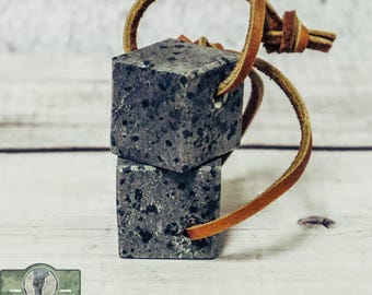 Puede incluir: Dos pequeños cubos de esteatita grises con motas negras están atados con un cordón de cuero marrón. Los cubos están apilados uno encima del otro. El fondo es una superficie de madera clara.