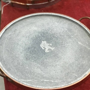 May include: A round, gray stone cooking surface with a copper rim and a small, stylized figure of a person in the center. The surface is slightly textured and has a circular pattern.