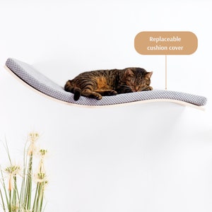 May include: A gray cat bed shaped like a curved shelf, mounted on a white wall. The bed has a gray fabric cushion with a replaceable cover. The cat is sleeping on the bed.