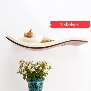 Può includere: Un letto bianco per gatti con un design curvo e un cuscino bianco morbido. Il letto è montato su una parete e ha una base in legno. Il testo "3 shelves" è scritto in rosso su uno sfondo bianco.