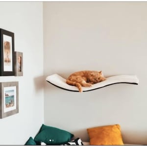 Puede incluir: Una cama para gatos blanca con forma de ola, montada en una pared, con un gato jengibre durmiendo en ella.