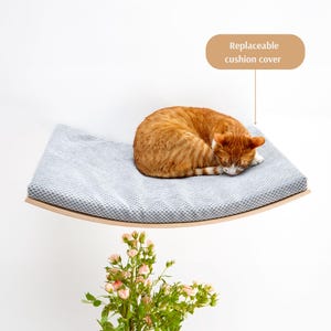 May include: A gray cat bed with a replaceable cushion cover, mounted on a wall. The bed is curved and made of wood. A ginger cat is sleeping on the bed.