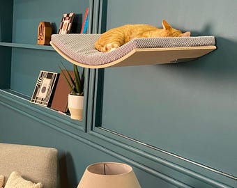 May include: A curved wooden cat shelf with a grey fabric cushion, mounted on a teal wall. A ginger cat is sleeping on the shelf.
