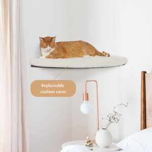 May include: A white, fluffy cat bed shelf mounted on a corner wall. The shelf is made of wood and has a curved shape. A cat is lying on the shelf, and the text "Replaceable cushion cover" is visible in the image.