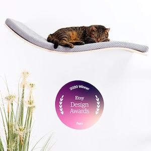Può includere: Un gatto tigrato marrone si riposa su una mensola curva grigia per gatti, fissata a una parete bianca. Un adesivo rotondo indica "2020 Winner Etsy Design Awards Pets".