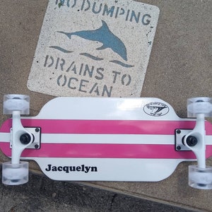 Könnte beinhalten: Weißes und rosafarbenes Skateboard mit schwarzen Trucks und transparenten Rollen. Das Deck hat einen weißen Streifen und die Worte "Jacquelyn" sind darauf gedruckt. Die Trucks haben schwarze Rollen mit transparentem Urethan.