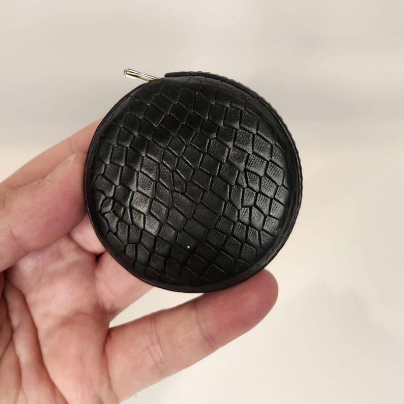 May include: A black, round tape measure with a textured, woven pattern. The tape measure has a metal pull tab and a small metal loop. The item is held in a hand against a white background.
