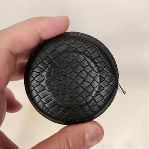 May include: A black, round measuring tape with a woven leather-like texture. The tape measure has a metal pull tab and is held in a hand. The tape measure is a useful tool for sewing, crafting, or home improvement projects.