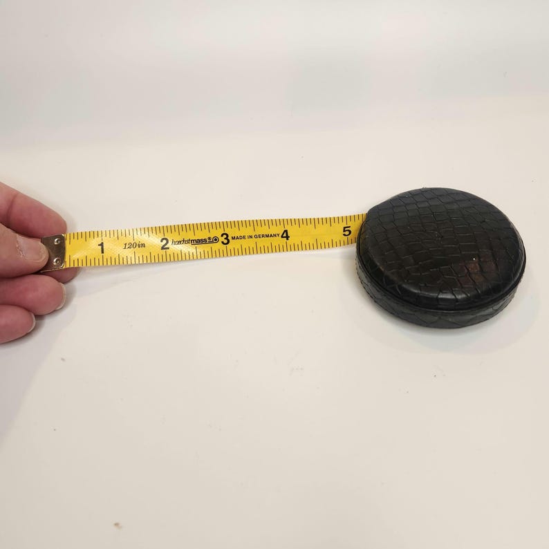 May include: A black, round tape measure with a textured surface. The yellow tape extends to 5 inches, with markings in inches and the text "Made in Germany". The tape measure is 120 inches long.