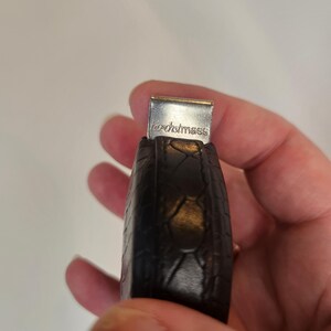 May include: A dark brown leather key fob with a textured pattern. The metal clasp at the top is silver and has the word "hochstmass" engraved on it. The key fob is held in a hand.