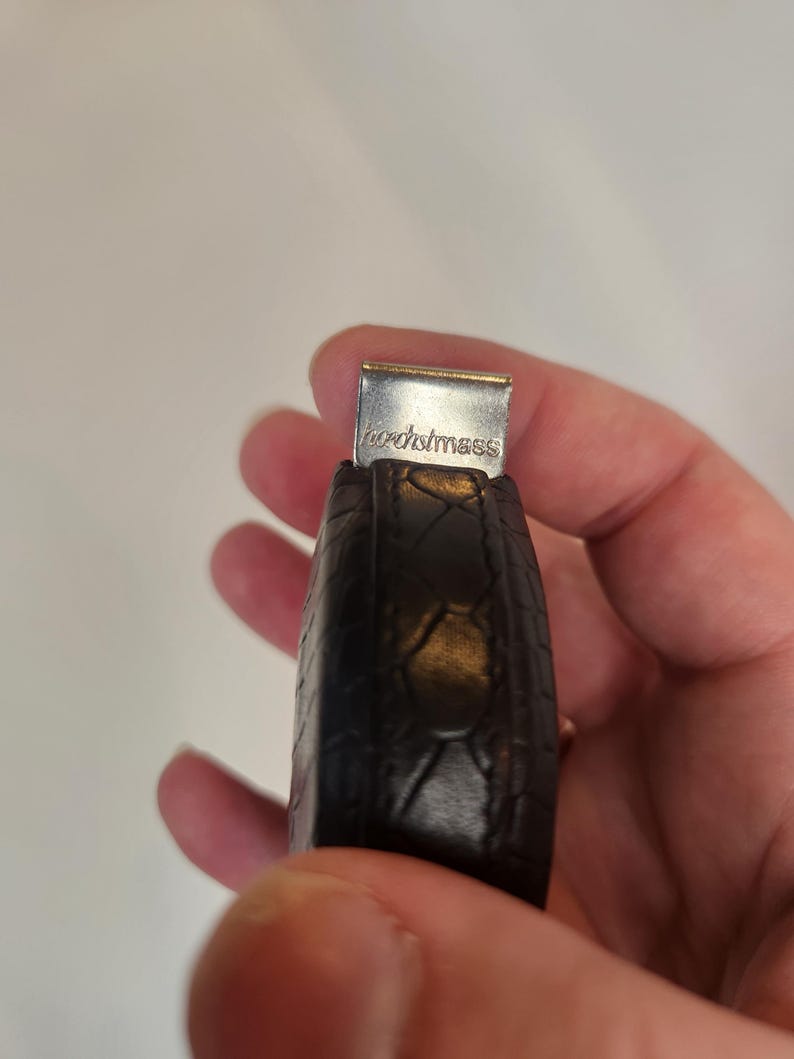 May include: A dark brown leather key fob with a textured pattern. The metal clasp at the top is silver and has the word "hochstmass" engraved on it. The key fob is held in a hand.