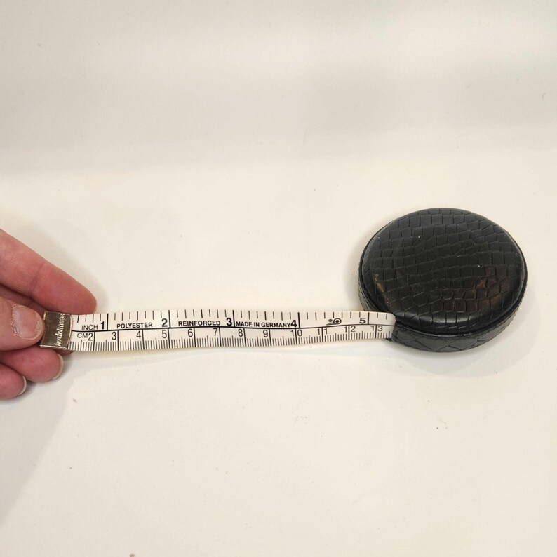 May include: A black, round tape measure with a textured surface. The white tape extends to 13 inches, marked with inch and centimeter measurements. The tape is labeled "Made in Germany."