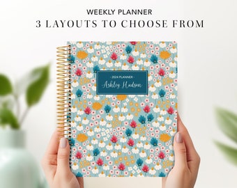 May include: A blue weekly planner with a floral pattern featuring white, yellow, pink, and blue flowers. The planner has a gold spiral binding and a personalized name tag that reads "2024 Planner - Ashley Hudson".