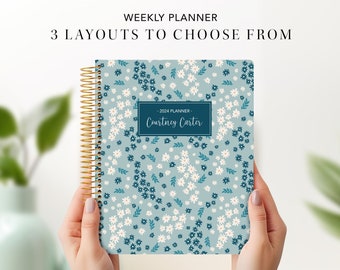 May include: A floral-patterned weekly planner with a gold spiral binding. The cover features a light blue background with white and dark blue flowers. Text on the cover reads "2024 Planner - Courtney Carter". The planner is held by two hands.